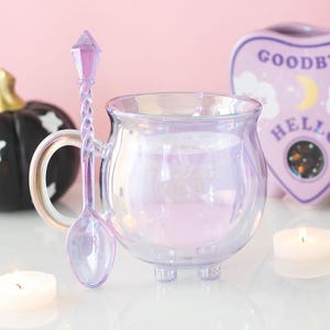 May include: A translucent, iridescent purple glass mug shaped like a small cauldron with three short feet and a curved handle. The mug includes a matching purple glass spoon with a twisted handle and a faceted, crystal-like top finial.