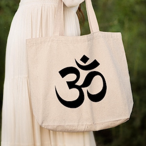 Może przedstawiać: Biała torba tote z płótna z nadrukowanym czarnym symbolem Om z przodu. Torba ma długie uchwyty i jest trzymana przez osobę w białej sukience.