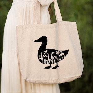 Może przedstawiać: Beżowa torba typu tote z czarną sylwetką kaczki z kwiatami wewnątrz sylwetki. Torba ma długi pasek i jest trzymana przez osobę.