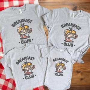 May include: Four grey t-shirts featuring a cartoon pancake design and the text "Breakfast Club". The shirts are in various sizes, with the largest on the left and the smallest on the right.