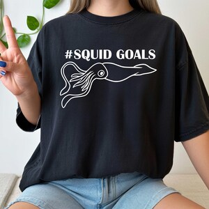 A person wearing a black t-shirt with a white graphic of an octopus and the text "#SQUID GOALS" printed on it.