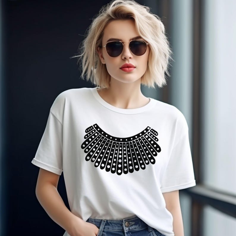 A woman with blonde hair wearing a white t-shirt with a black graphic design on it, standing in front of a window.