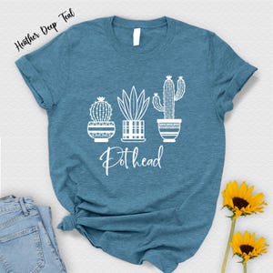 May include: A heather deep teal t-shirt with a white graphic of three potted plants. The plants are a cactus, a succulent, and a leafy plant. The text "Pothead" is printed below the plants.