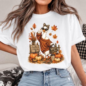May include: A white t-shirt featuring a graphic of a goose wearing a brown sweater, leopard print headband, and sunglasses. The goose holds a leopard print handbag. The scene includes orange autumn leaves, pumpkins, and a sign reading hello FALL.