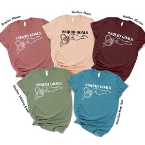 a collection of five t-shirts, each with a unique color and design, featuring the text "# SQUID GOALS" printed on them.