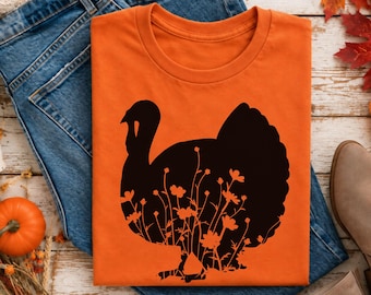 May include: An orange short-sleeved t-shirt featuring a black silhouette of a turkey. The turkey graphic is filled with a detailed pattern of various wildflowers and stems, creating a contrast against the solid orange fabric of the shirt.