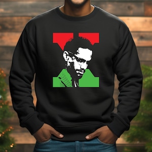 May include: Black sweatshirt with a graphic portrait in white, green, and red. The design includes a red 'T' shape above the portrait and a green rectangle below. The man is wearing glasses and a green jacket.