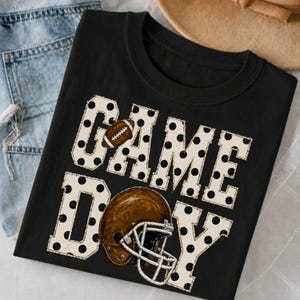Könnte beinhalten: Ein schwarzes T-Shirt mit Rundhalsausschnitt und dem Schriftzug GAME DAY in großen, cremefarbenen Blockbuchstaben mit schwarzen Punkten. Ein brauner Football liegt auf dem Buchstaben A und ein brauner Footballhelm mit weißem Gitter ist darunter.