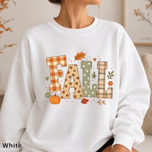 Puede incluir: Una sudadera blanca de manga larga con la palabra FALL en letras grandes. Cada letra tiene un patrón diferente: cuadros vichy naranjas, flores, lunares verdes y tartán. El diseño incluye pequeñas calabazas naranjas y hojas.