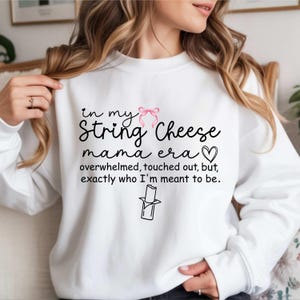 Könnte beinhalten: Weißes Sweatshirt mit dem Text "in my String Cheese mama era" in schwarzer Schrift, einer rosa Schleife und einem Herz. Der Text geht weiter mit "overwhelmed, touched out, but, exactly who I'm meant to be." Eine Grafik von Käsefäden ist ebenfalls enthalten.