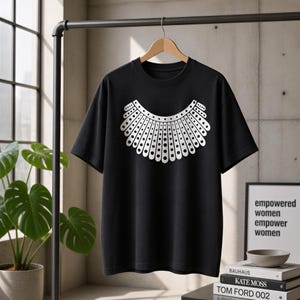 A black t-shirt with a white design of a rooster's head is hanging on a clothing rack in a room with a potted plant and books.