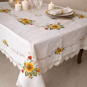 May include: White linen tablecloth featuring embroidered sunflowers and a delicate lace trim. The table is set with candles, glassware, and a floral arrangement. The text "THE LINEN PETAL & Handcrafted Lace Tablecloth" is visible.