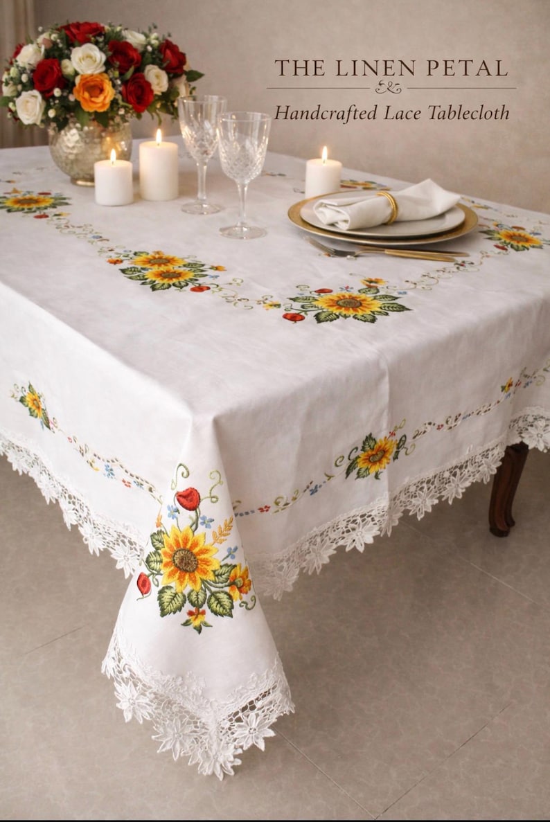 May include: White linen tablecloth featuring embroidered sunflowers and a delicate lace trim. The table is set with candles, glassware, and a floral arrangement. The text "THE LINEN PETAL & Handcrafted Lace Tablecloth" is visible.