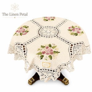 Puede incluir: Un mantel redondo de color crema con bordes de encaje de ganchillo intrincados y cuatro grupos florales bordados en tonos rosa, verde y amarillo. Una etiqueta blanca rectangular con el texto The Linen Petal está adherida.
