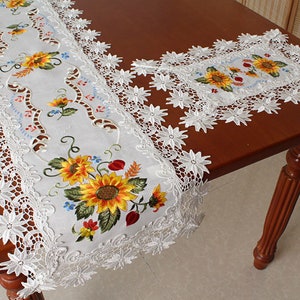 May include: White embroidered table runner and matching placemat with a sunflower design. The runner has a scalloped lace edge. The sunflowers are yellow with green leaves and red accents. The placemat is square.
