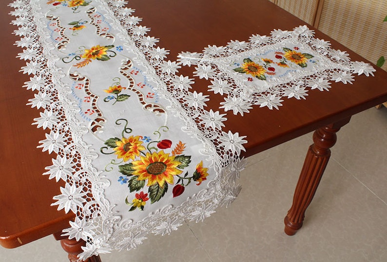 May include: White embroidered table runner and matching placemat with a sunflower design. The runner has a scalloped lace edge. The sunflowers are yellow with green leaves and red accents. The placemat is square.
