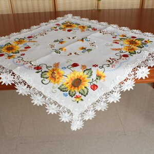 May include: White square tablecloth featuring embroidered sunflowers in yellow, orange, and green. The tablecloth has a scalloped lace edge. The sunflowers are the focal point of the design, with detailed petals and centres.