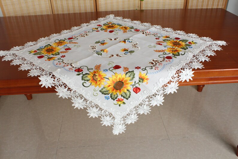 May include: White square tablecloth featuring embroidered sunflowers in yellow, orange, and green. The tablecloth has a scalloped lace edge. The sunflowers are the focal point of the design, with detailed petals and centres.