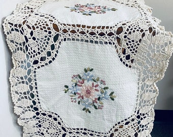 May include: White crocheted tablecloth with embroidered floral designs. The tablecloth has a square shape with rounded corners and a delicate crocheted border.