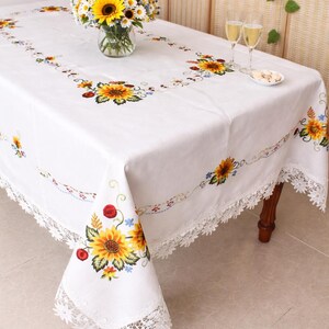 May include: White rectangular tablecloth with embroidered sunflowers, red berries, and blue flowers. The tablecloth has a delicate lace trim. A vase of sunflowers and daisies sits on the table.