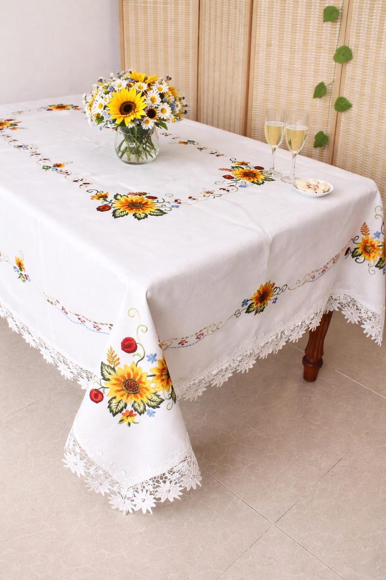 May include: White rectangular tablecloth with embroidered sunflowers, red berries, and blue flowers. The tablecloth has a delicate lace trim. A vase of sunflowers and daisies sits on the table.
