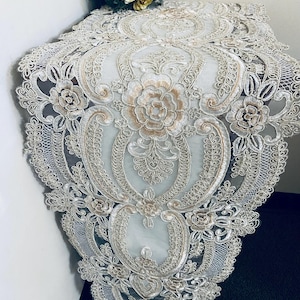 May include: Cream-colored lace table runner with intricate floral and scrollwork embroidery. The runner features a central floral medallion and delicate scalloped edges. A decorative piece for dining tables or other surfaces.
