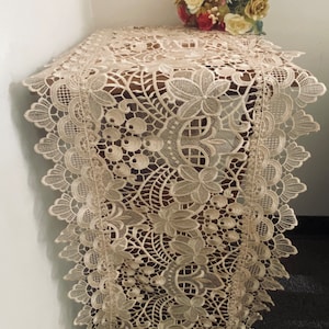 May include: A beige lace table runner with a delicate floral pattern. The runner has a scalloped edge and is made of a lightweight, airy fabric.