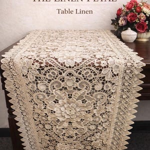 May include: Beige lace table runner with a floral design. The runner is displayed on a dark brown table. The text "THE LINEN PETAL Table Linen" is at the top of the image. A vase of flowers and a small white container are on the table.