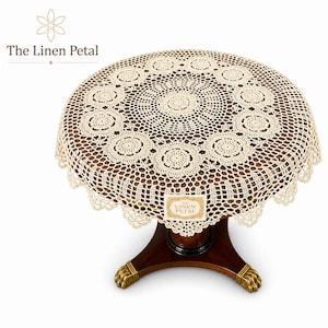 May include: A cream-colored, crocheted table topper with a floral design, covering a round, dark wooden table. The table has a pedestal base with gold-colored claw feet. The Linen Petal logo is visible.