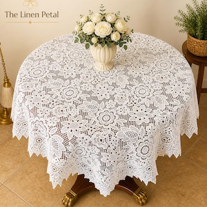 May include: A round white lace tablecloth with a floral pattern covers a wooden table. A vase of white roses sits in the center. The tablecloth has a scalloped edge. The Linen Petal is printed in the top left corner.