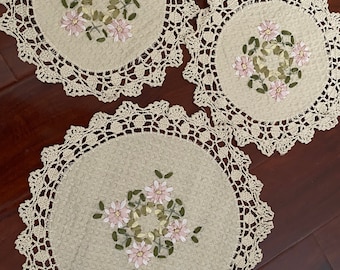 May include: Three crocheted doilies with delicate floral embroidery in pink and green. The doilies are made of a beige cotton yarn and have a scalloped edge.