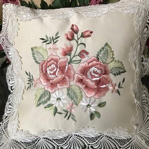 May include: Cream-colored square pillow with embroidered roses in shades of pink and green. The pillow is trimmed with white lace. The floral design includes pink buds, white daisies, and green leaves. The pillow is a decorative home accent.