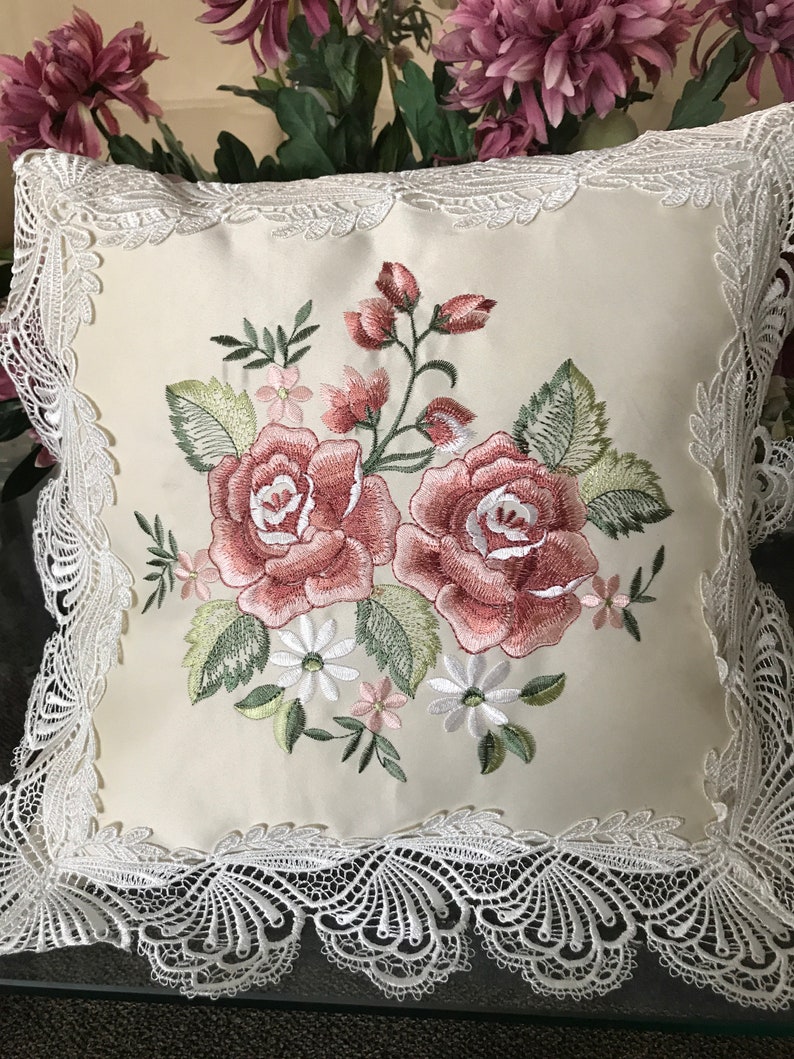 May include: Cream-colored square pillow with embroidered roses in shades of pink and green. The pillow is trimmed with white lace. The floral design includes pink buds, white daisies, and green leaves. The pillow is a decorative home accent.