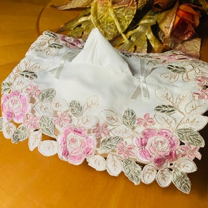 May include: White tissue box cover with delicate pink and green floral embroidery. The cover features a lace-like design with a raised center for the tissue box.