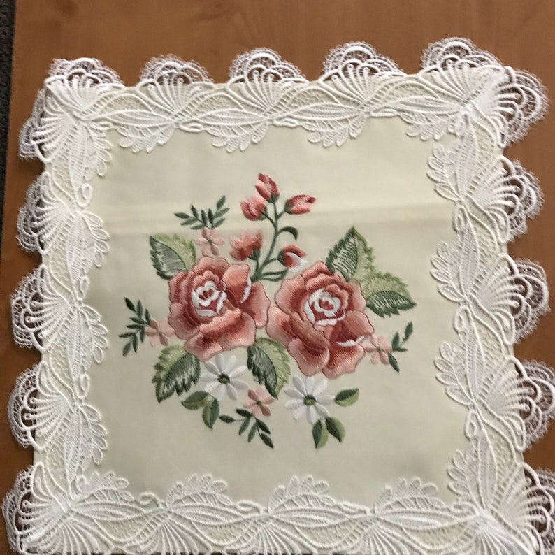 May include: Cream-colored square table runner with an embroidered floral design. The embroidery features two large pink roses, smaller pink buds, white daisies, and green leaves. The edges are trimmed with a white lace border.