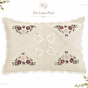 May include: Cream-colored rectangular pillow with a crocheted design and embroidered floral accents. The pillow features a central pattern and four corner designs with pink and burgundy flowers and green leaves. The text "The Linen Petal" is at the top.