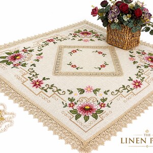 May include: Cream-colored embroidered tablecloth with a floral design featuring pink and green flowers. The tablecloth has a square shape with a lace trim and a central embroidered square. A basket of flowers and the text "THE LINEN PETAL" are also visible.