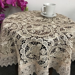 May include: A beige lace table runner with a delicate floral pattern. The runner is round and has a scalloped edge. It is placed on a wooden table with a white cup and saucer on top.