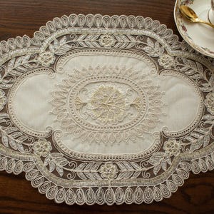 May include: Oval-shaped ivory lace table runner with intricate floral and leaf patterns. The runner features a detailed, embroidered design with a scalloped edge. A teacup and saucer with a gold spoon are visible in the upper right corner.