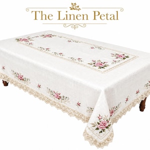 May include: Cream-colored rectangular tablecloth with embroidered pink roses and green leaves. The tablecloth has a decorative lace trim and the text "The Linen Petal" in gold.