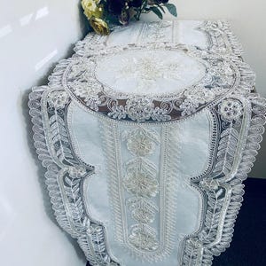 May include: White lace table runner with floral embroidery and scalloped edges. The runner features an oval centerpiece and detailed floral patterns. A bouquet of flowers sits on the table.