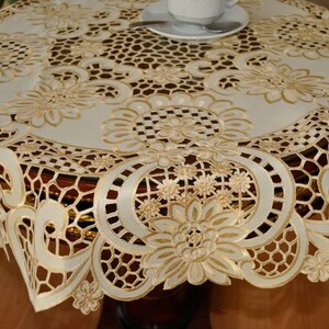 May include: Cream-colored round tablecloth with intricate floral embroidery and cutwork details. The tablecloth is draped over a dark wooden table, with a white teacup and saucer visible on top. The design features a repeating floral pattern.