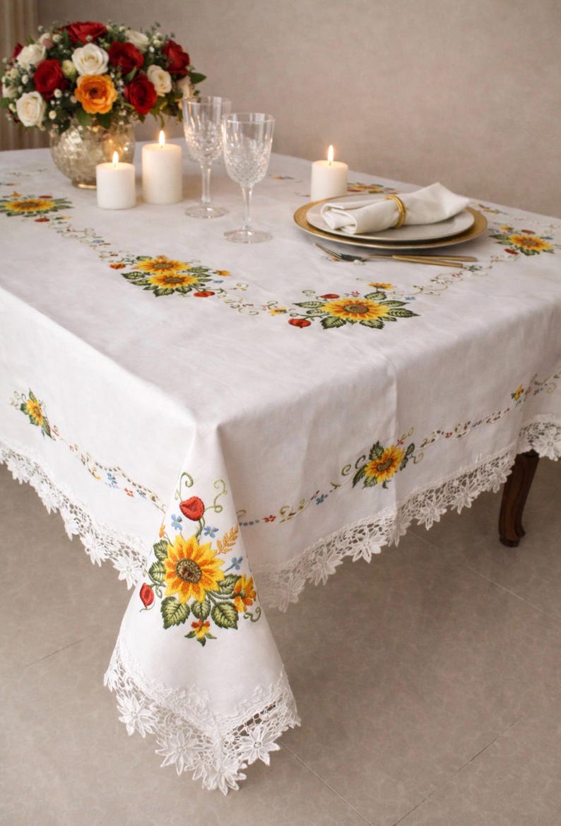 May include: White rectangular tablecloth with embroidered sunflowers, red poppies, and green leaves. The tablecloth has a delicate lace trim and is set with candles, glassware, and a floral centerpiece, creating an elegant dining setting.