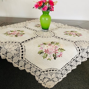 May include: A white crocheted tablecloth with a delicate floral embroidery design. The tablecloth features a square shape with rounded corners and a lace border. A vase with pink flowers sits on top of the tablecloth.