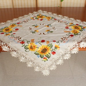 May include: A square beige tablecloth featuring embroidered sunflowers, red berries, and green leaves. The tablecloth has a scalloped lace edge. The sunflowers are yellow and orange. The tablecloth is on a wooden table.