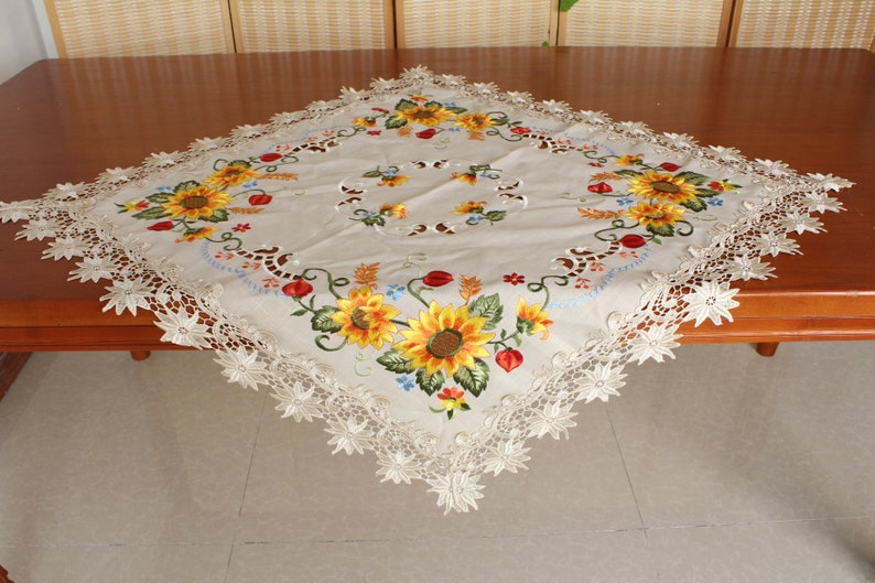 May include: A square beige tablecloth featuring embroidered sunflowers, red berries, and green leaves. The tablecloth has a scalloped lace edge. The sunflowers are yellow and orange. The tablecloth is on a wooden table.