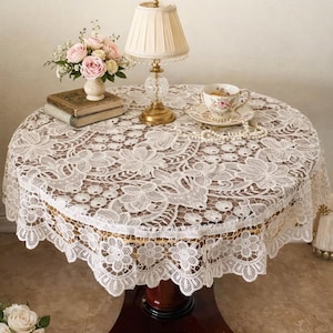 May include: A round, vintage lace table topper with a floral design, set on a dark wood table. The table is adorned with a lamp, a teacup, a vase of pink roses, and a stack of books. The text "The Linen Petal" is at the top.