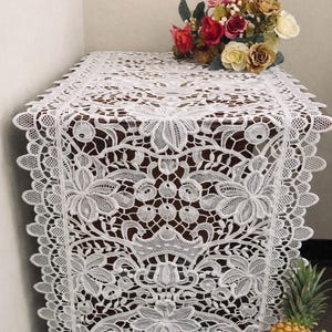 May include: White lace table runner with intricate floral and leaf patterns. The runner is displayed on a dark wooden table, with a bouquet of roses and other flowers on top. The text "THE LINEN PETAL Table Linen" is at the top.