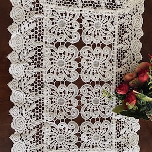 May include: A white lace table runner with a floral design. The runner features a repeating pattern of flowers and leaves, with a scalloped edge. The runner is rectangular and appears to be made of cotton or a similar material.