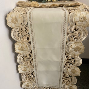 May include: Cream-colored table runner with intricate floral embroidery and a scalloped edge. The runner is placed on a table, with a bouquet of flowers in the background. The design features a central panel and decorative borders.